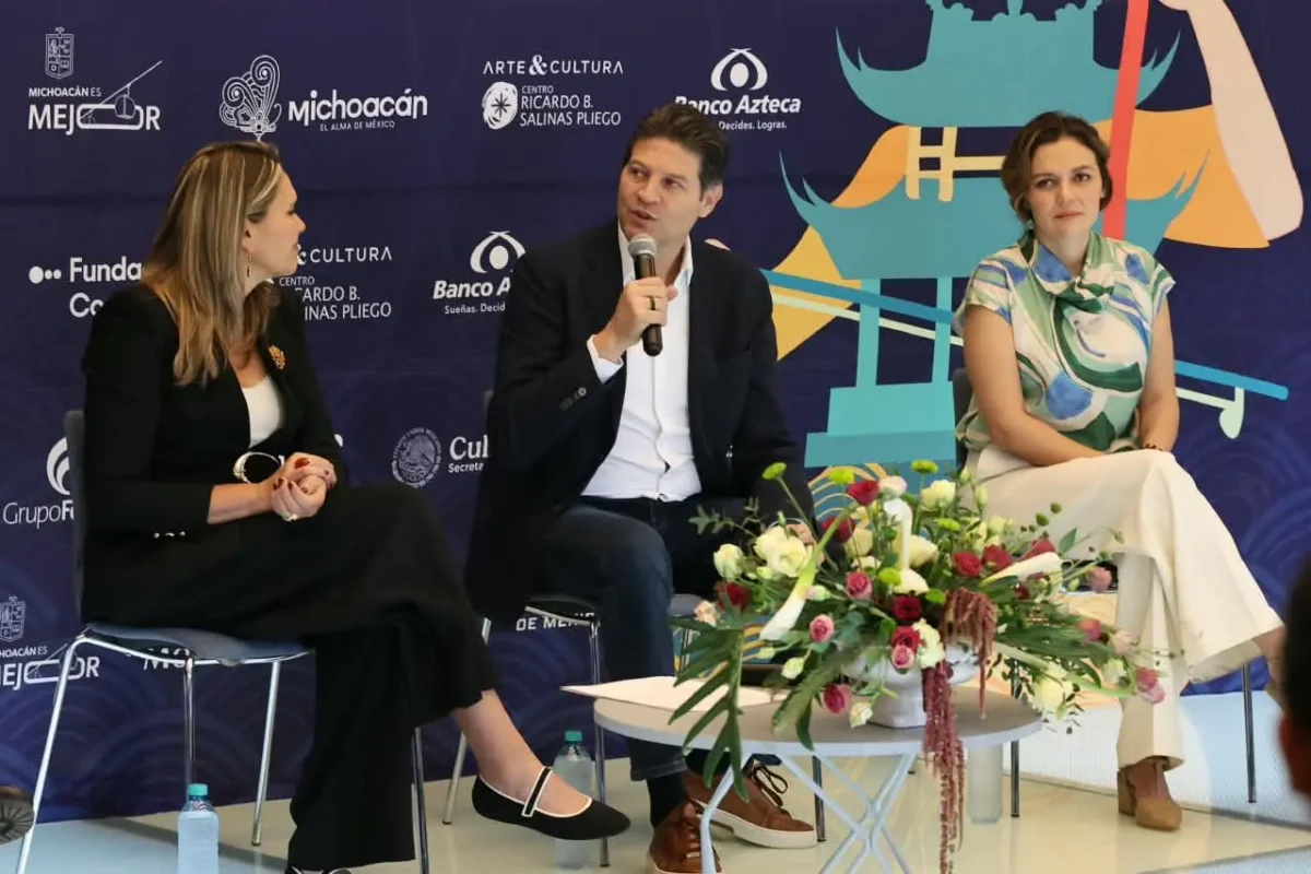 Tres personas en una mesa durante una conferencia, con fondo de logotipos y arte cultural.