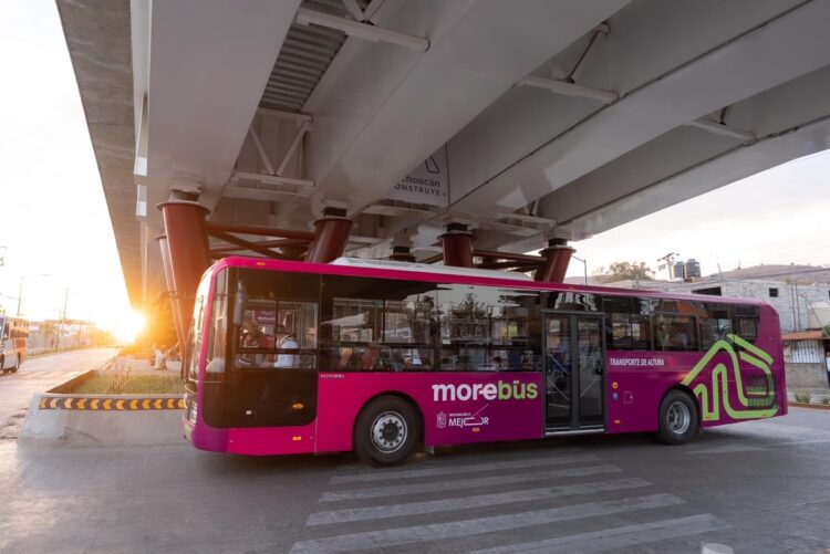 Autobús rosa de transporte público en Michoacán, bajo un puente, con sol en el horizonte y fondo urbano.