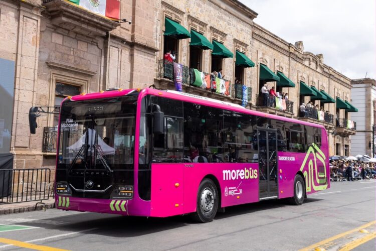 Un autobús urbano rosa circula por una calle junto a un edificio histórico con balcones y personas observando desde las ventanas.