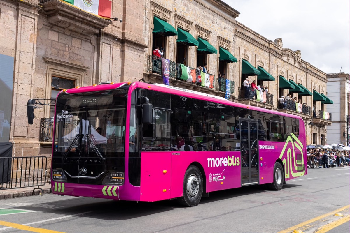 Un autobús urbano rosa circula por una calle junto a un edificio histórico con balcones y personas observando desde las ventanas.