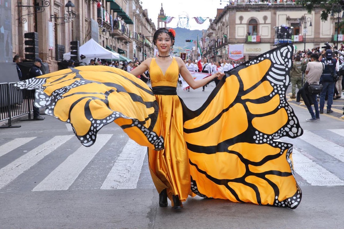 Una mujer con un vestido amarillo que imita las alas de una mariposa camina en un desfile en la calle.
