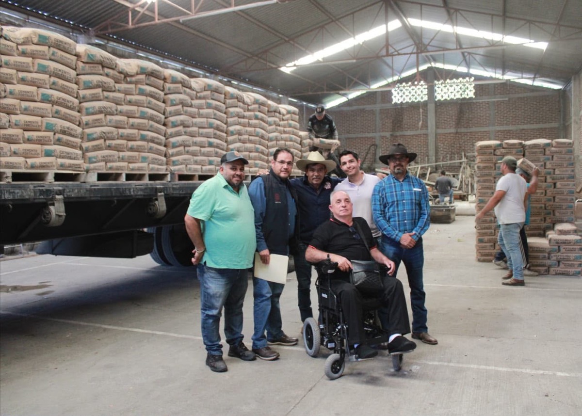 Grupo de personas en almacén de cemento con sacos apilados y maquinaria en segundo plano.