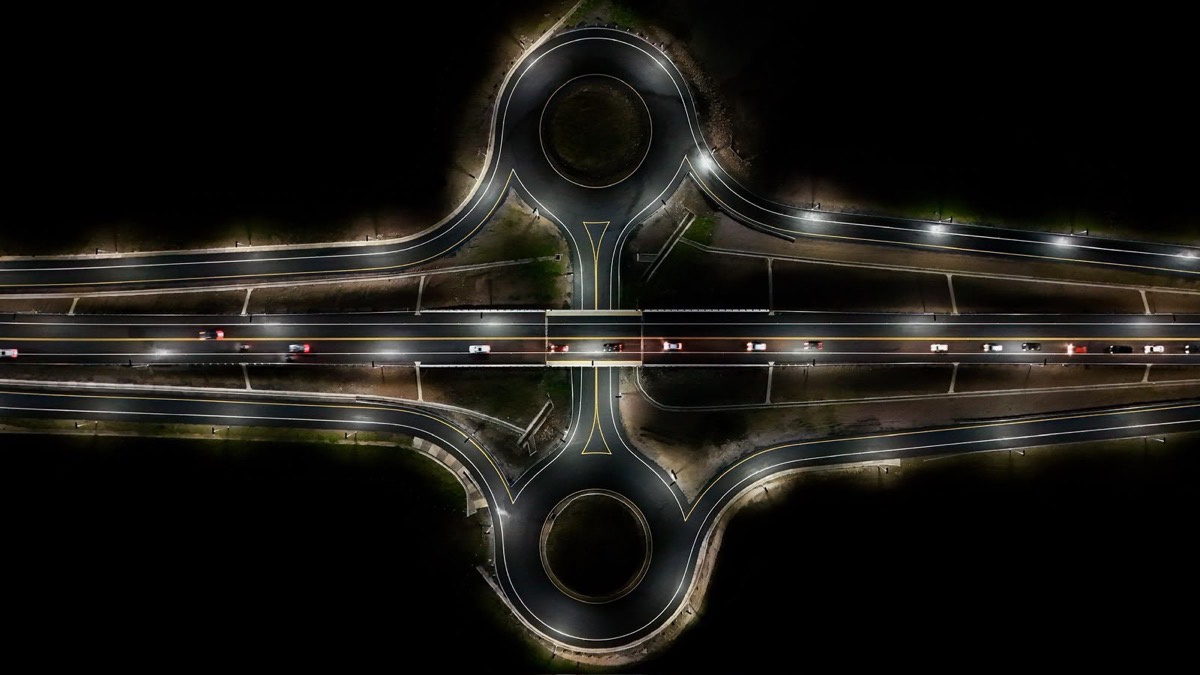 Vista aérea de una rotonda con vías de circunvalación iluminadas en la noche, mostrando movimiento vehicular.