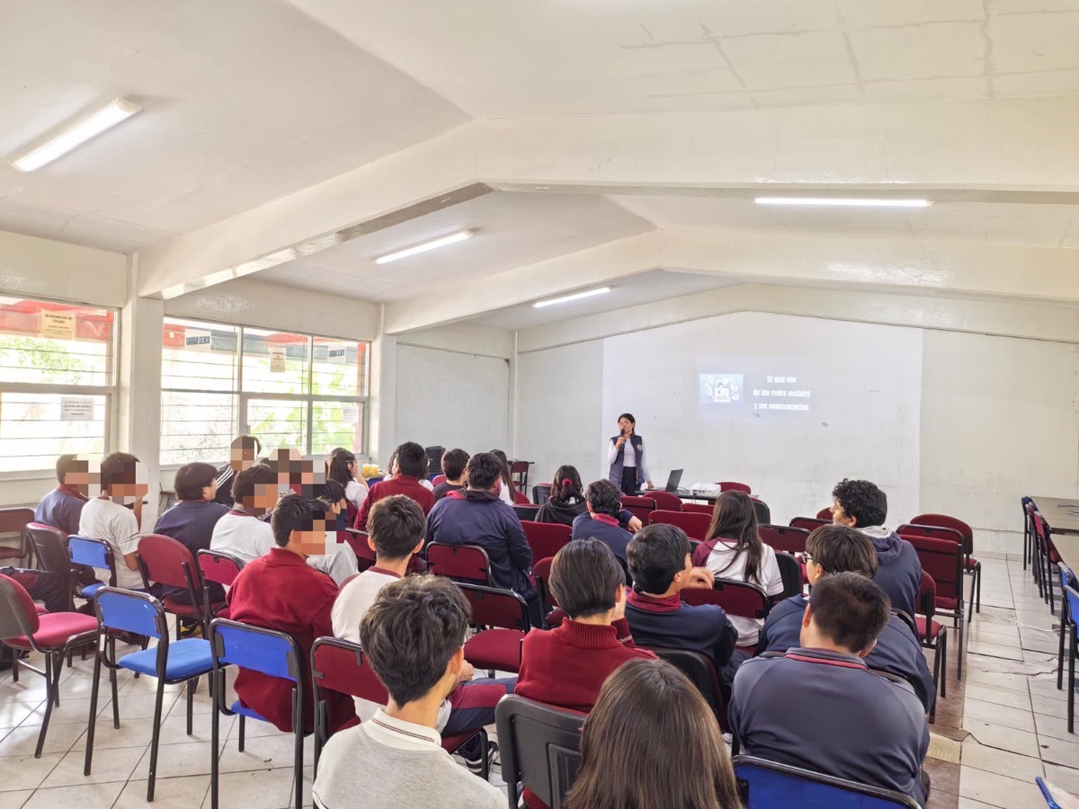 Un grupo de estudiantes está sentado en un salón mientras una persona da una presentación proyectada en la pared.
