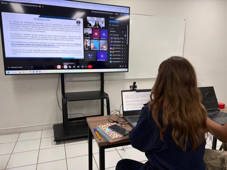 Estudiante participando en clase virtual con pantalla grande y computadora portátil en aula moderna.