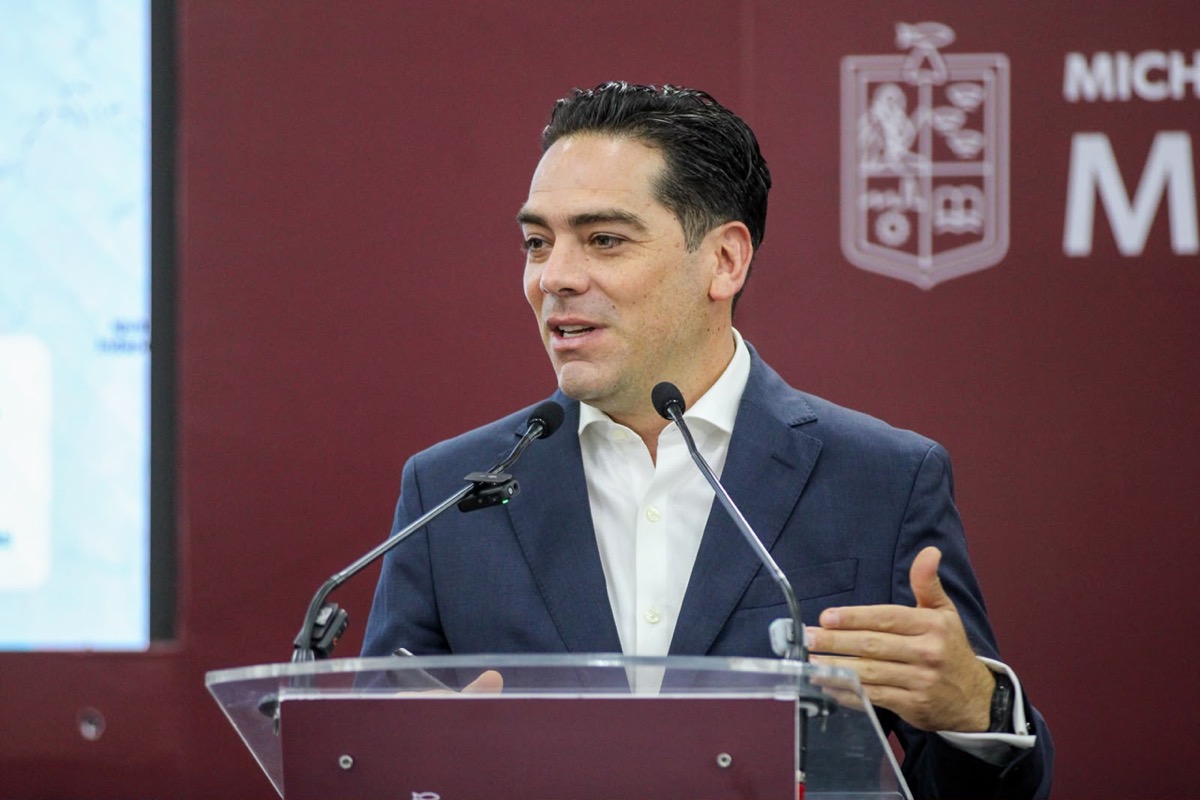Hombre en traje azul y camisa blanca da un discurso en una conferencia con micrófonos y fondo rojo con logotipo de Michoacán.