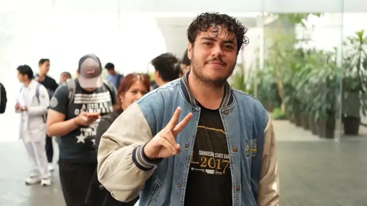 Joven con chaqueta y camiseta en evento público, haciendo señal de paz con dos dedos, en un ambiente con muchas personas y植物.
