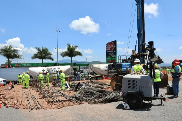 Trabajadores y maquinaria en construcción del distribuidor vial en Zamora, con cielo despejado y palmeras en el fondo.