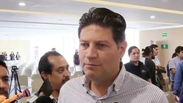 Alfonso Martínez en evento público con fondo de asistentes y cámaras en un espacio cerrado.