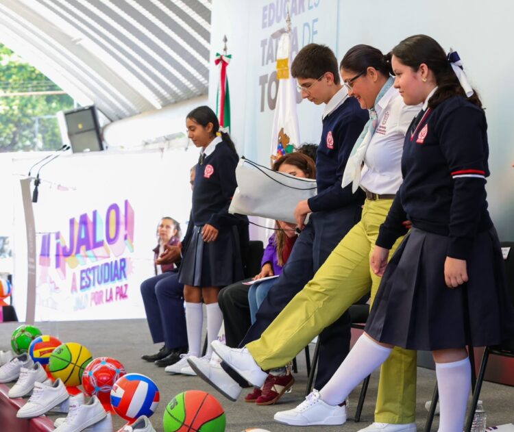 Un grupo de estudiantes y una adulta patean pelotas coloridas en un escenario durante un evento escolar.