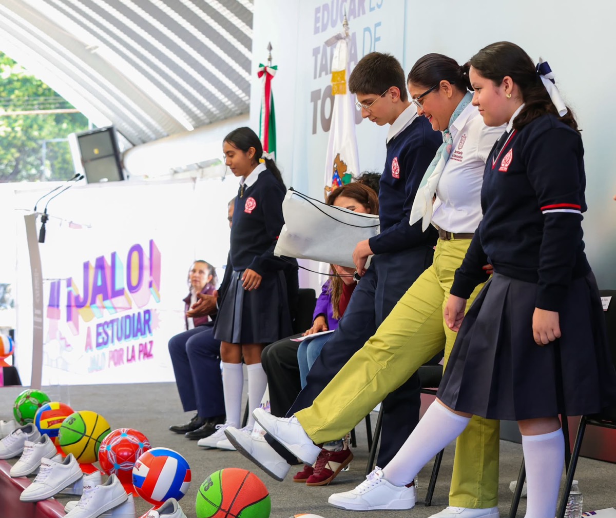 Un grupo de estudiantes y una adulta patean pelotas coloridas en un escenario durante un evento escolar.