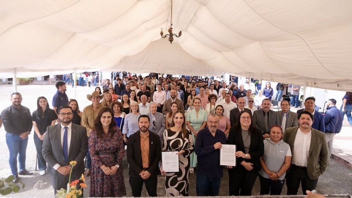 Un grupo grande de personas posa bajo una carpa blanca durante un evento, con varias personas al frente sosteniendo documentos.