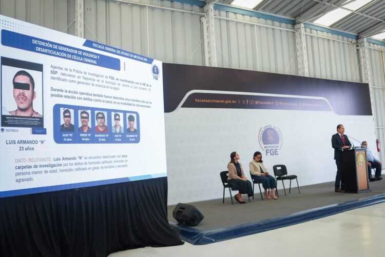 Un hombre da una conferencia en un podio junto a una pantalla que muestra fotos y datos de personas detenidas, mientras varias personas están sentadas en el escenario.