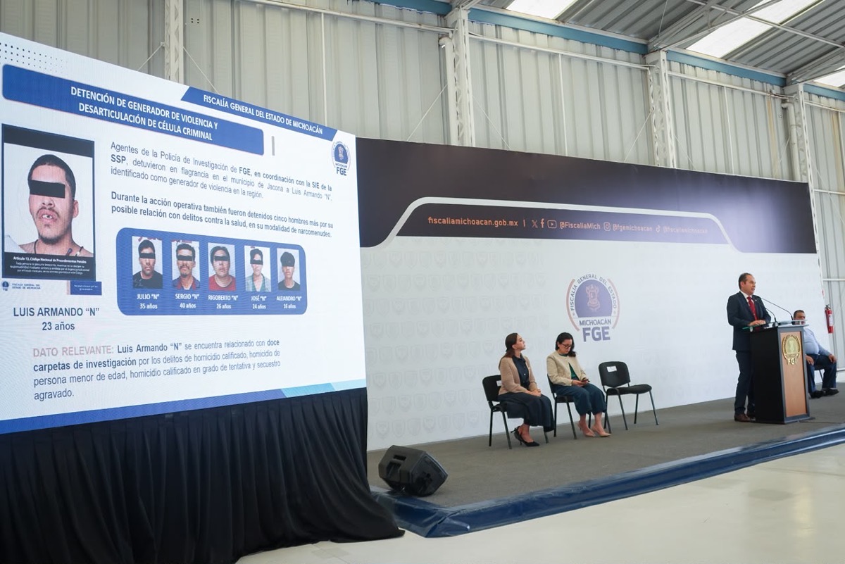Un hombre da una conferencia en un podio junto a una pantalla que muestra fotos y datos de personas detenidas, mientras varias personas están sentadas en el escenario.