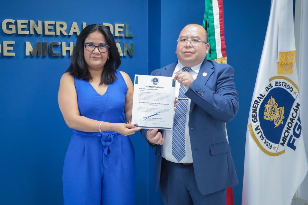Dos personas posan sosteniendo un documento frente a una pared azul con un logotipo oficial y una bandera a un costado.
