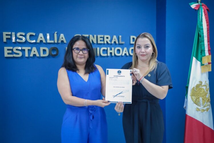 Dos mujeres posan frente a una pared azul mientras sostienen un documento, junto a una bandera de México.