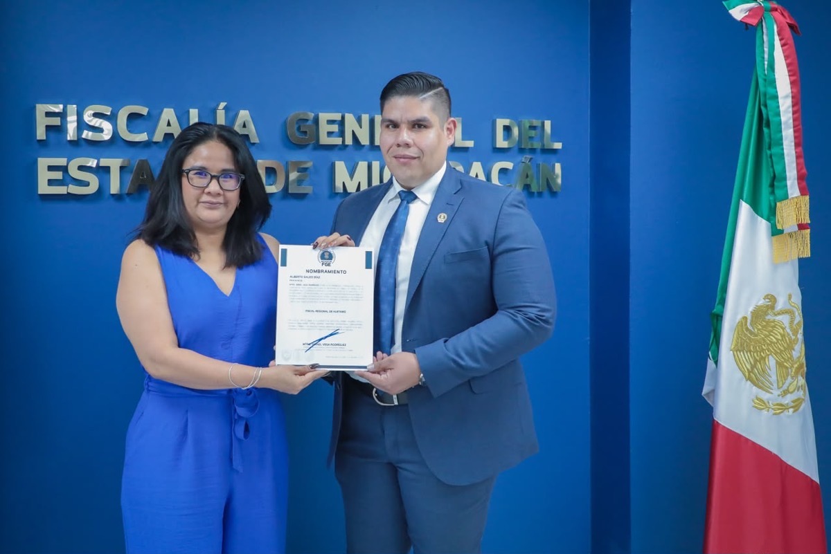 Dos personas posan frente a una pared azul sosteniendo un documento, junto a una bandera de México.