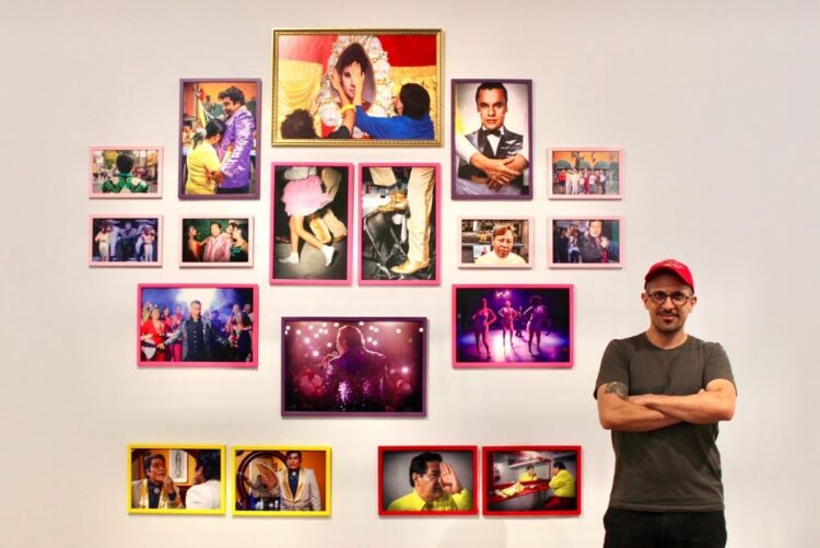 Un hombre con gorra roja está de pie frente a una pared con varias fotos coloridas enmarcadas.