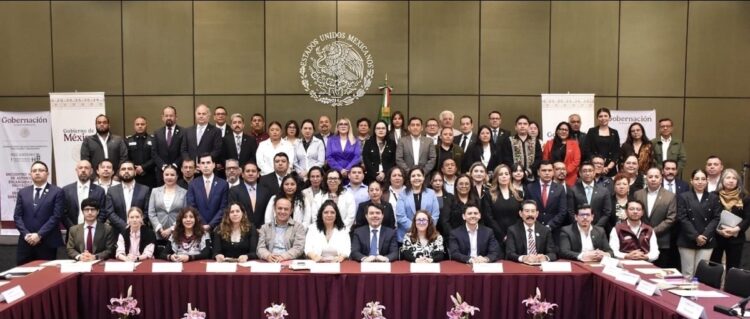 Un gran grupo de personas posa para una foto oficial en una sala con un emblema de México en la pared.