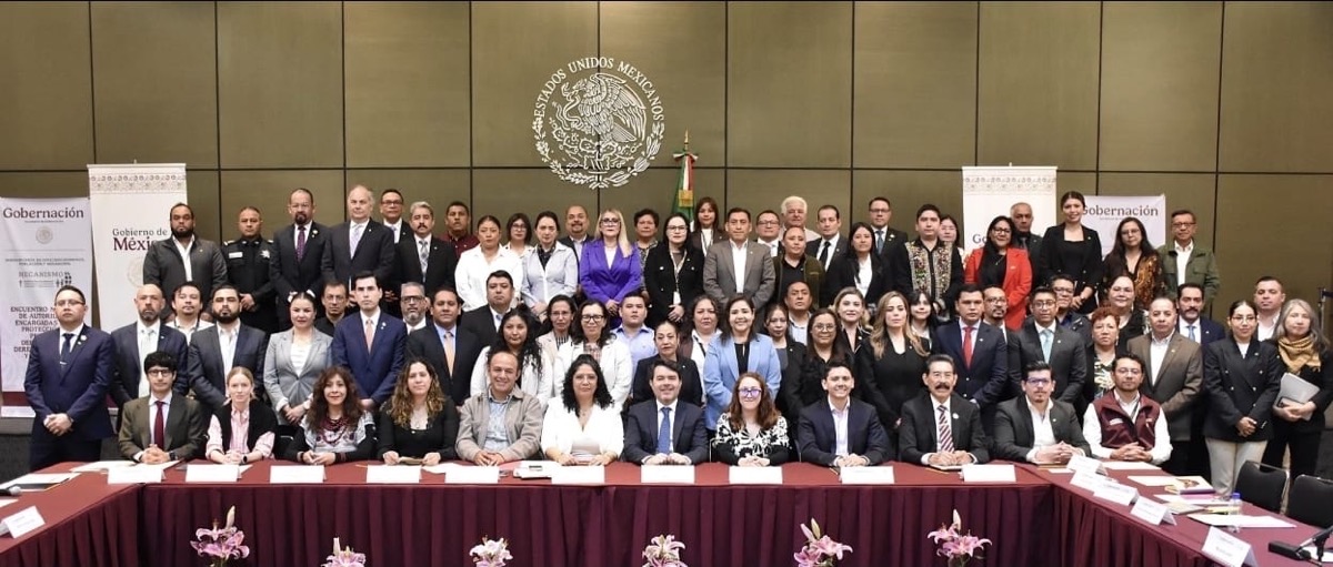 Un gran grupo de personas posa para una foto oficial en una sala con un emblema de México en la pared.