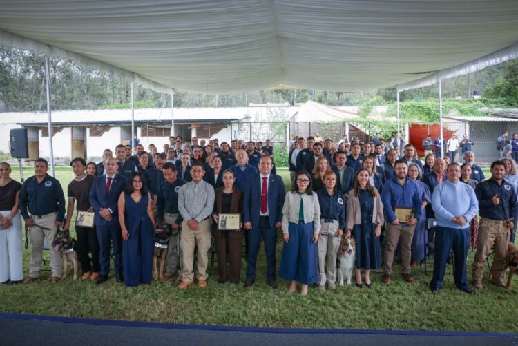 Un grupo grande de personas posa para una foto bajo una carpa al aire libre, algunas sosteniendo diplomas y acompañadas de perros.