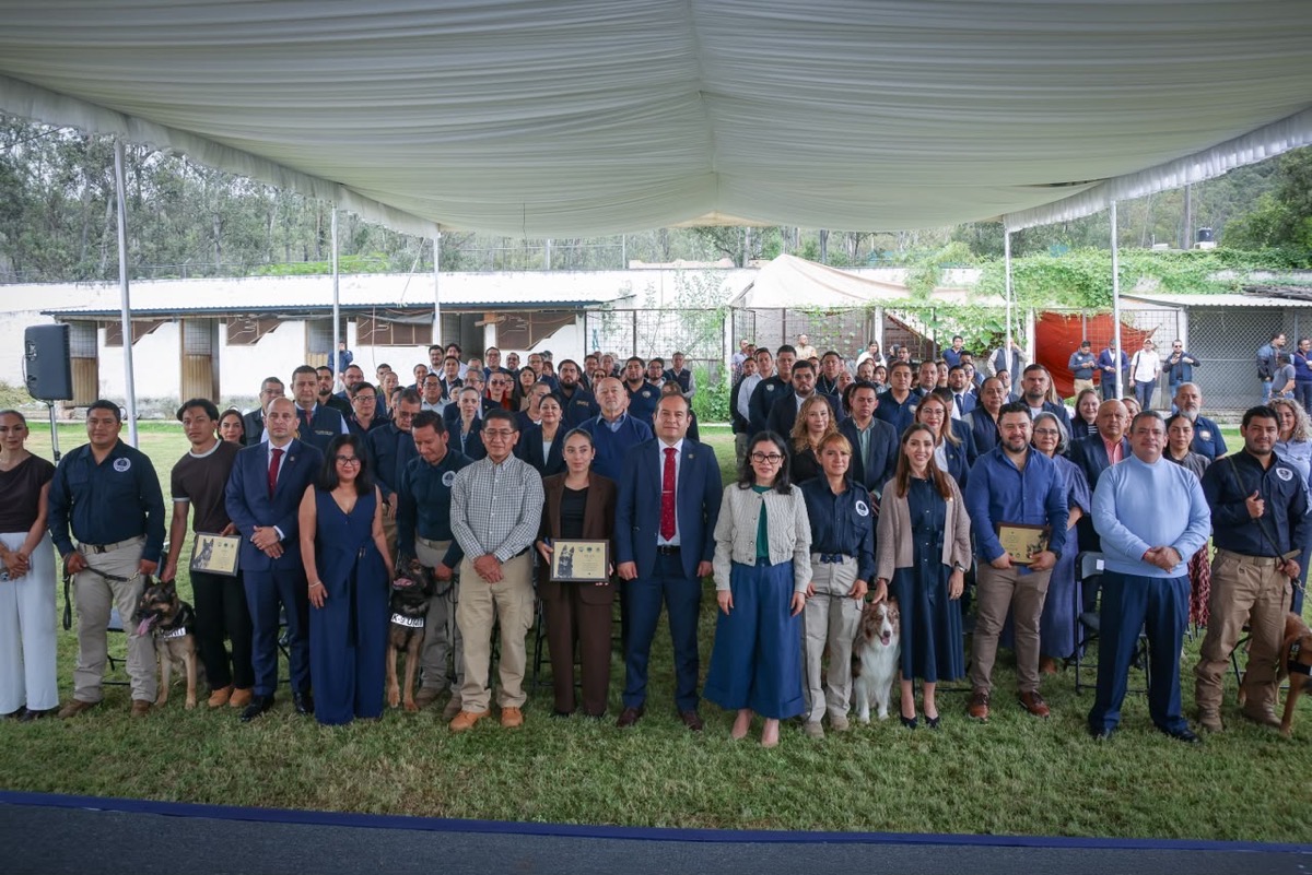 Un grupo grande de personas posa para una foto bajo una carpa al aire libre, algunas sosteniendo diplomas y acompañadas de perros.