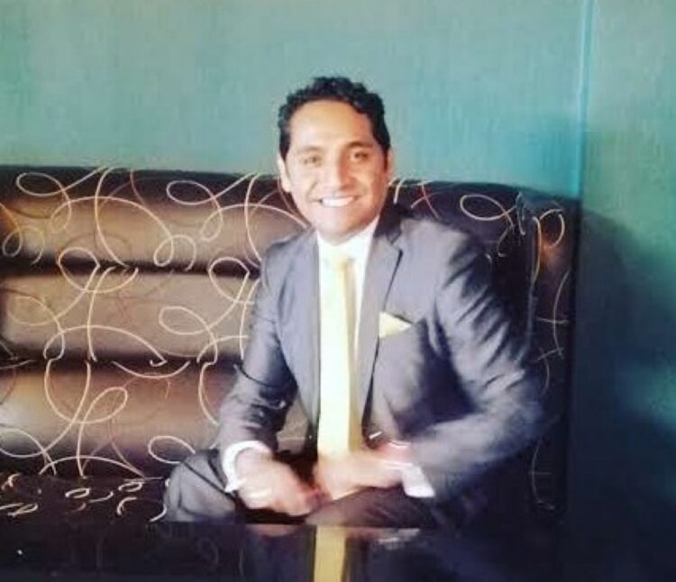 Tayde González en traje gris y corbata amarilla sentado en un sillón con fondo azul.
