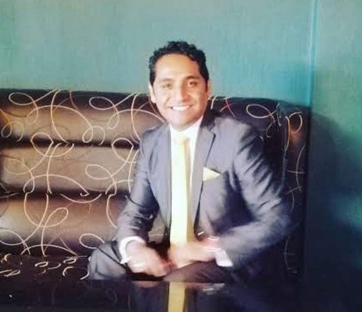 Tayde González en traje gris y corbata amarilla sentado en un sillón con fondo azul.