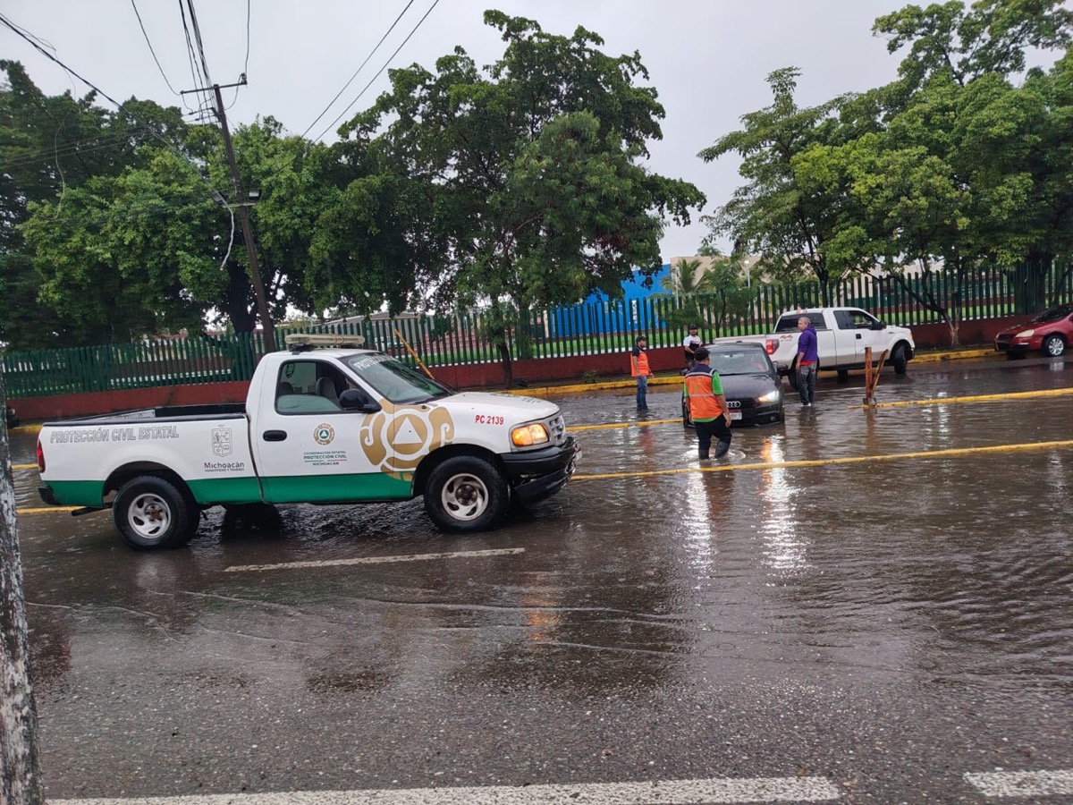 Una calle mojada con vehículos y personas, incluyendo trabajadores de protección civil, durante un día lluvioso.