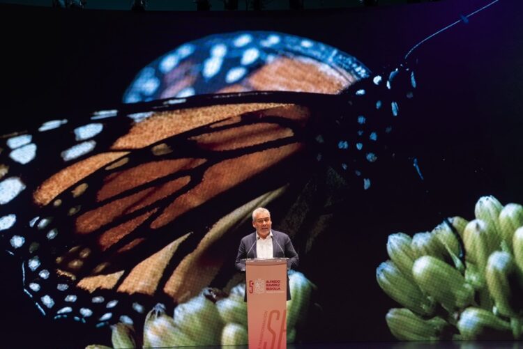 Un hombre habla en un podio frente a una pantalla gigante que muestra una mariposa monarca sobre flores verdes.