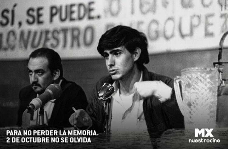 Dos hombres están sentados en una mesa con micrófonos, uno de ellos hablando y sosteniendo un cigarro, frente a un cartel parcialmente visible.