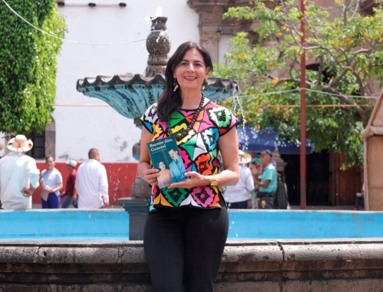 Mujer sonriente sosteniendo un libro en una plaza con fuente y árboles verdes, en Michoacán.