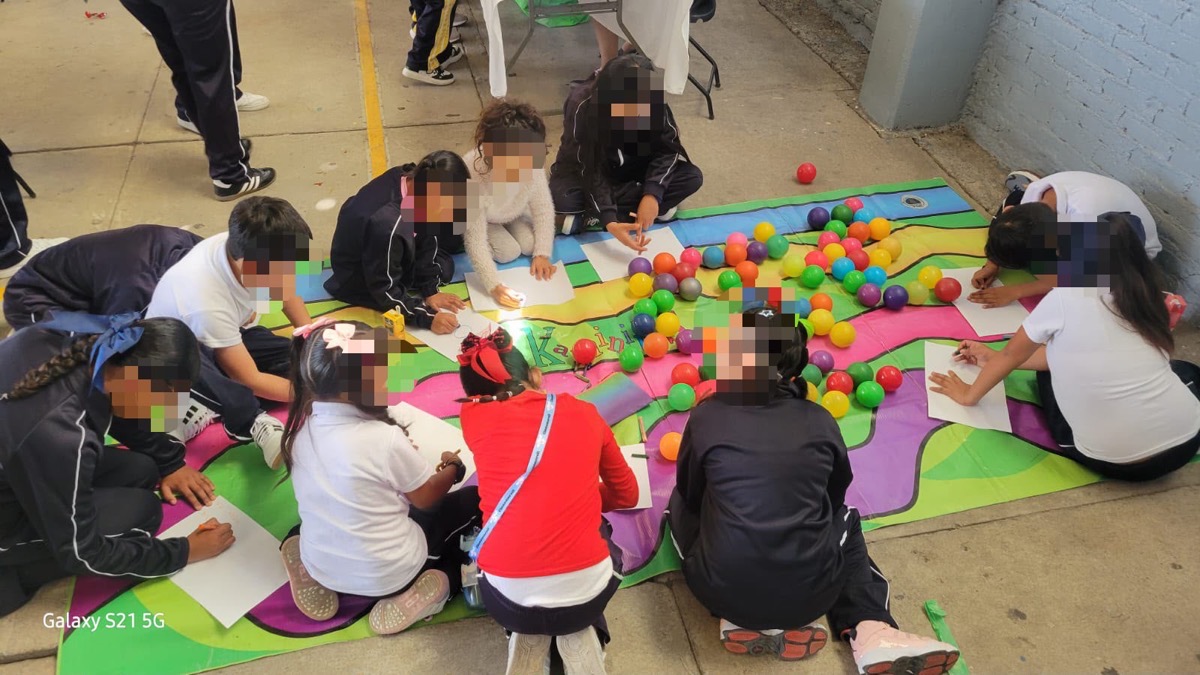 Niños y adultos en actividad lúdica con pelotas de colores en aula escolar, algunos escriben en cuadernos.