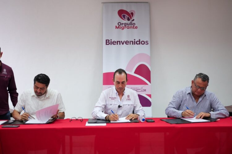Tres hombres sentados en una mesa roja firman documentos frente a un cartel que dice “Orgullo Migrante Bienvenidos”.