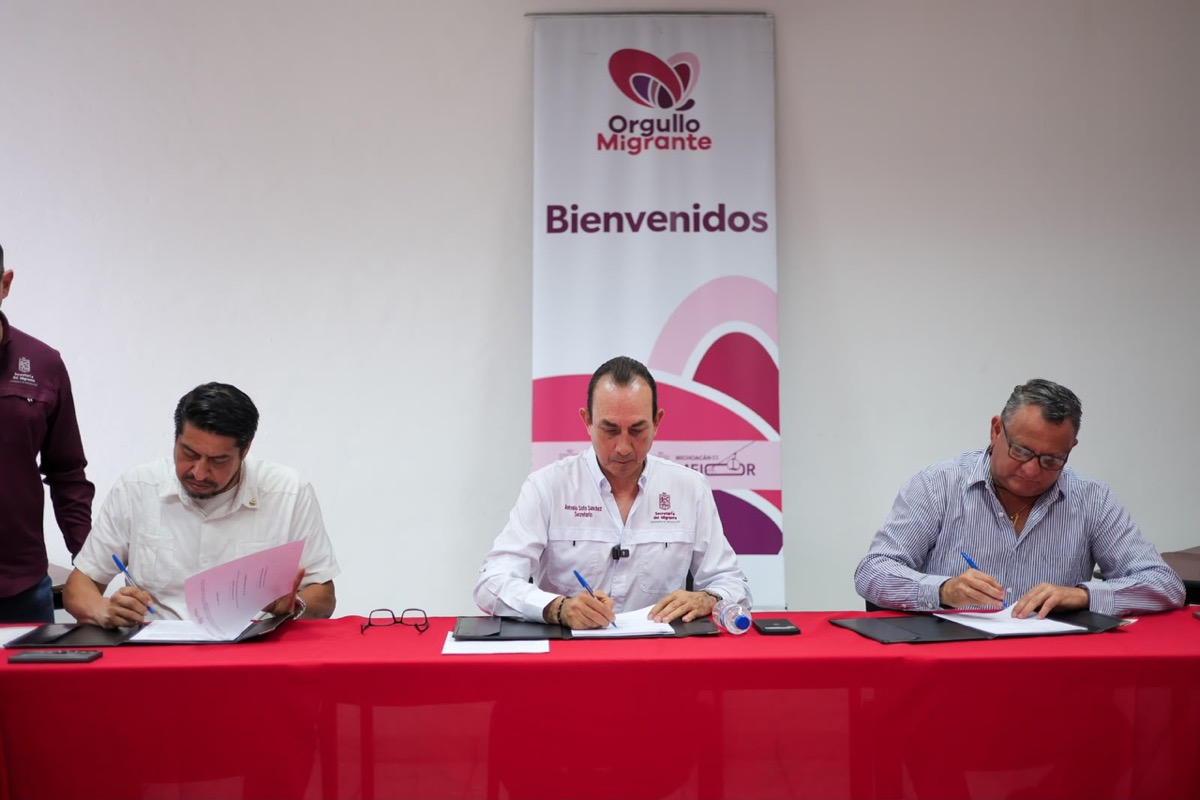 Tres hombres sentados en una mesa roja firman documentos frente a un cartel que dice “Orgullo Migrante Bienvenidos”.