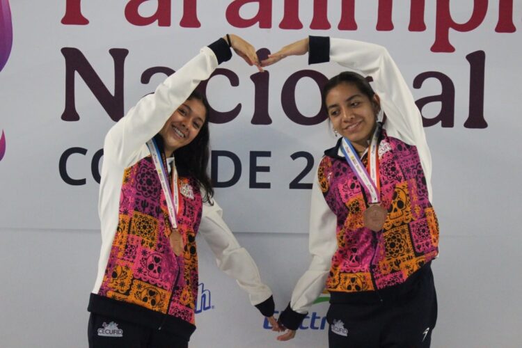 Dos jóvenes deportistas mexicanas con medallas en el cuello, formando un corazón con sus manos en un evento deportivo.