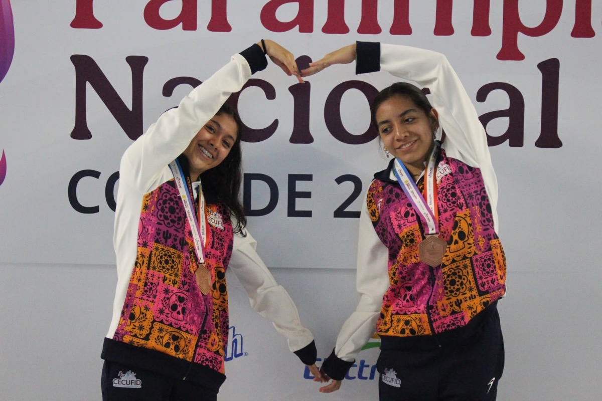 Dos jóvenes deportistas mexicanas con medallas en el cuello, formando un corazón con sus manos en un evento deportivo.