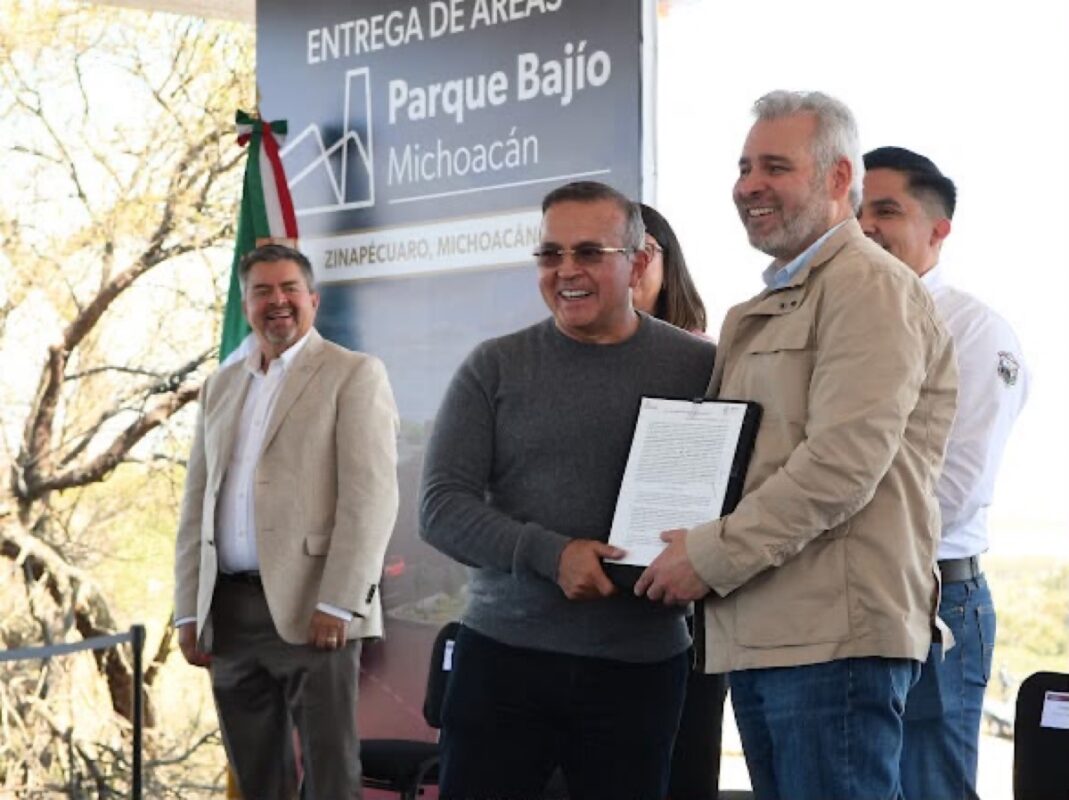Autoridades entregan documento en evento en Parque Bajo, Michoacán, con fondo de árboles y bandera mexicana.