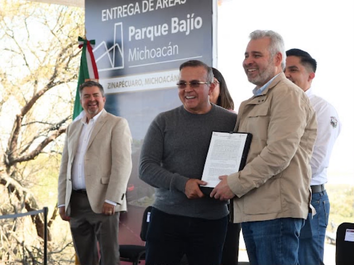 Autoridades entregan documento en evento en Parque Bajo, Michoacán, con fondo de árboles y bandera mexicana.