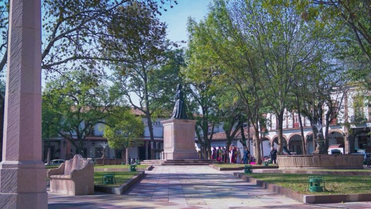 Una plaza arbolada con una estatua central, una fuente y personas caminando alrededor.