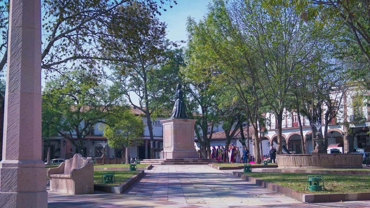 Una plaza arbolada con una estatua central, una fuente y personas caminando alrededor.