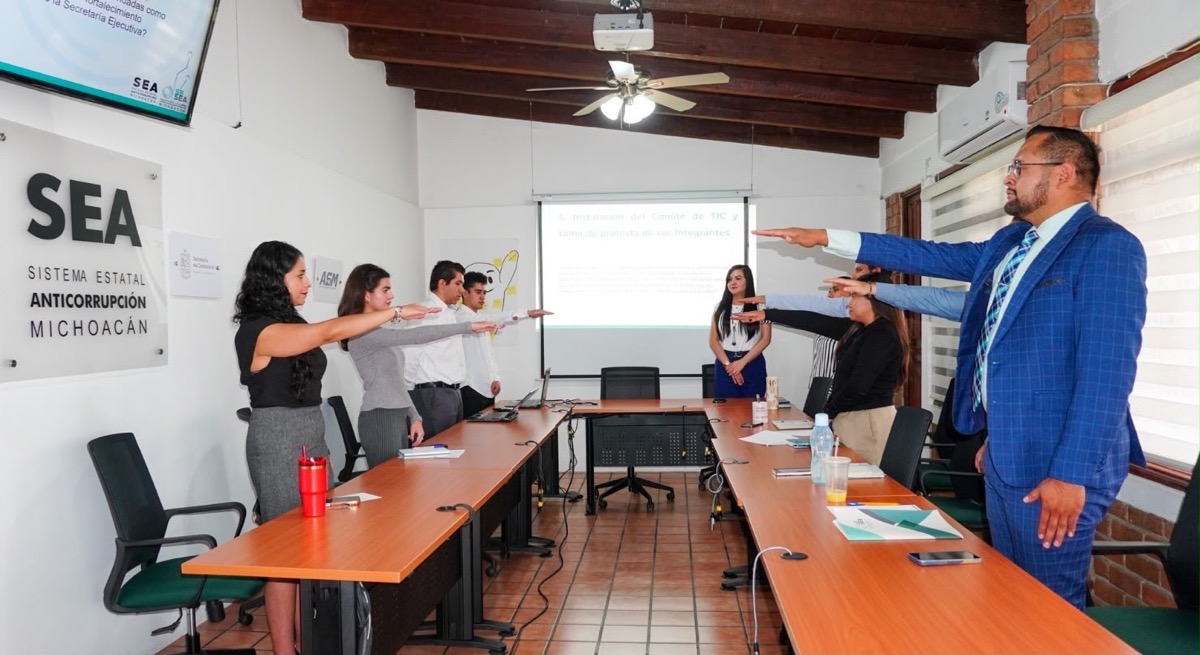 Un grupo de personas en una sala de reuniones levanta el brazo derecho mientras una mujer de pie los observa frente a una pantalla de proyección.