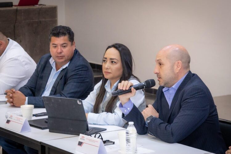 Tres personas sentadas en una mesa participan en una reunión, y una de ellas habla por un micrófono.