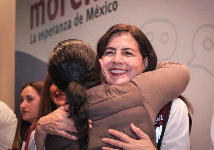 Una mujer sonriente abraza a otra persona en un evento mientras otras personas observan al fondo.