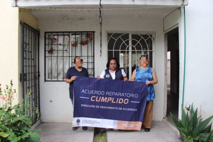 Tres personas están de pie frente a una casa sosteniendo una lona que dice “Acuerdo reparatorio cumplido”.