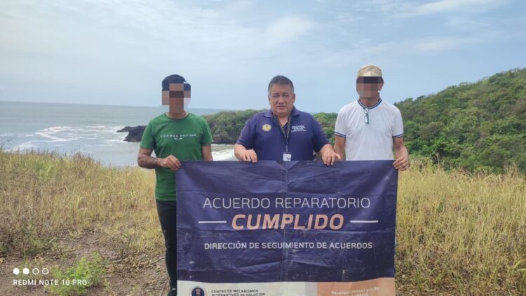 Tres hombres sostienen una lona que dice “Acuerdo Reparatorio Cumplido” en un paisaje costero con vegetación y el mar al fondo.