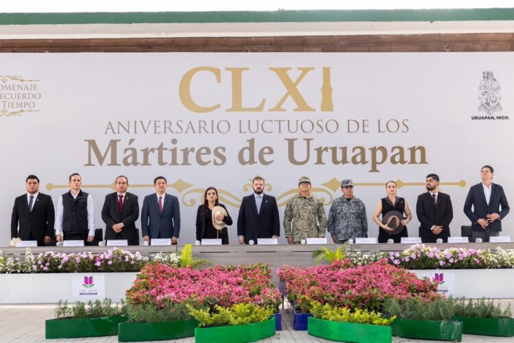 Un grupo de personas, incluidos funcionarios y militares, están de pie en un escenario decorado con flores durante una ceremonia con un cartel que dice “CLXI Aniversario Luctuoso de los Mártires de Uruapan”.