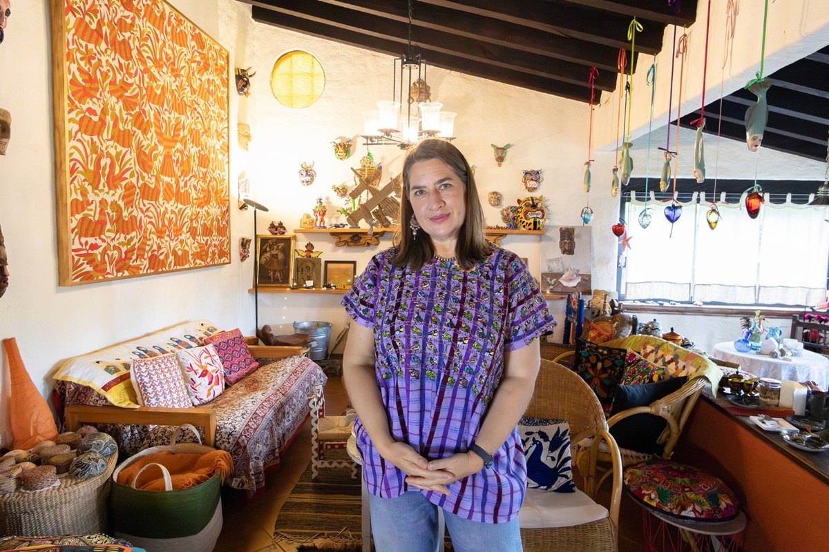 Una mujer de pie sonríe en una sala colorida decorada con artesanías, cuadros y cojines bordados.