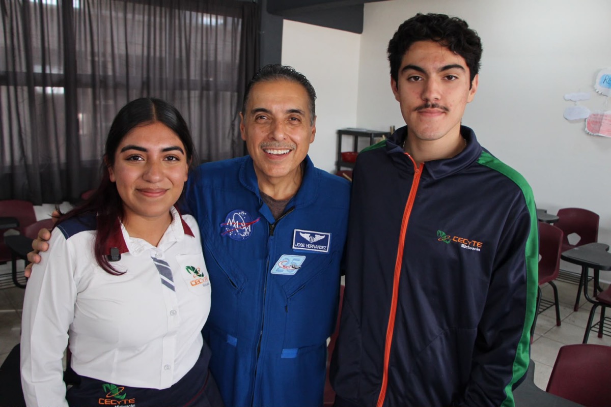 Tres personas sonríen mientras posan juntas en un aula, dos con uniformes de CECyTE y una con un traje azul de la NASA.