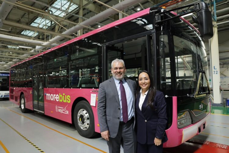 Dos personas de pie y sonriendo frente a un autobús rosa dentro de una nave industrial.